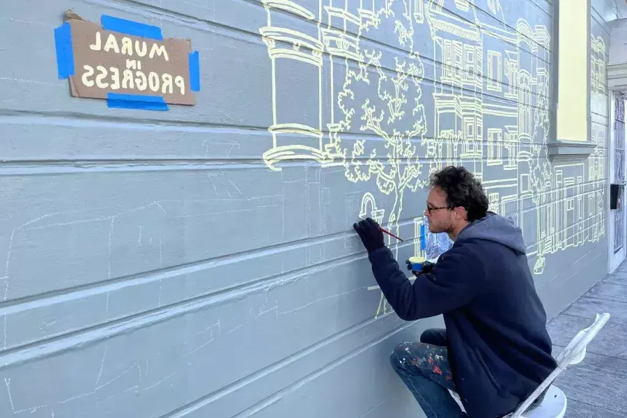 An artist paints a mural on the side of a building in the Mission District, with a sign taped onto the building that reads "Mural in Progress.San Francisco, CA.